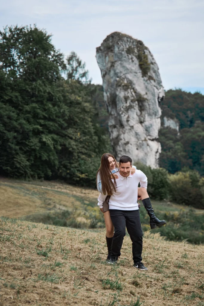 mezczyzna niesie na plecach usmiechnieta kobiete na trawiastym zboczu u podnoza skaly